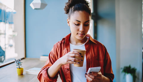 A young Black woman drinks a hot beverage while looking at her cell phone.